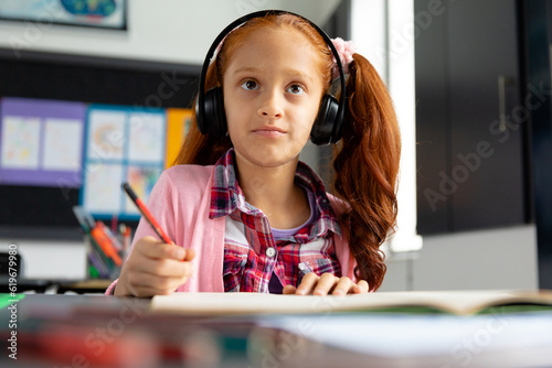 Happy biracial schoolgirl sitting at desk wearing headphones in class