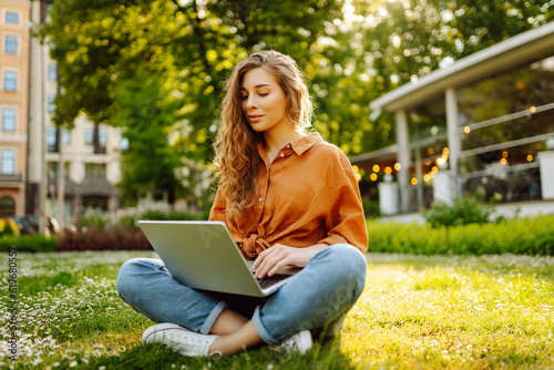 Beautiful female freelancer sits on a green meadow with a laptop. Online education. Happy woman running outdoors enjoying the sun. Freelancing concept. © maxbelchenko