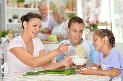 Portrait of cute brother and sister, mother eating together in kitchen