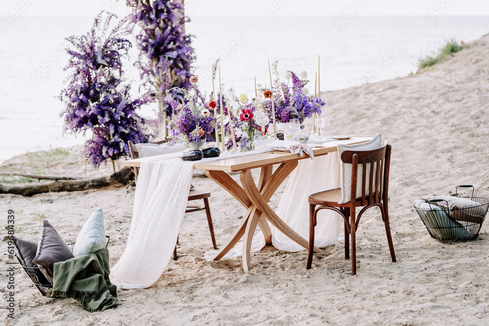 Fototapeta premium Dining Table and Chairs on Sandy Beach Nature Photography. Desk Decorated Natural Fresh Flowers and Plant for Celebrate Valentine Day on Seashore. Picnic Date for Lovely Couple Outside