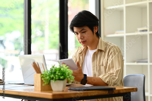 Handsome young businessman reading book or checking his working schedule plan while sitting at desk