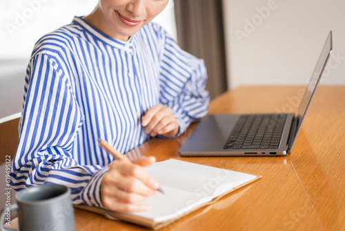 自宅でノートパソコンを開きノートに書いている女性 photo