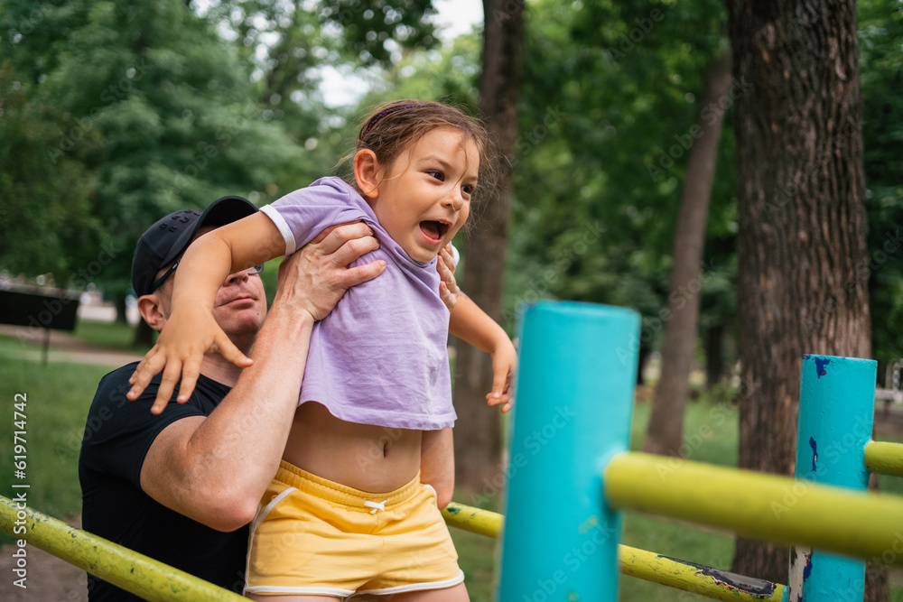 Father with child girl doing pull-ups on workout outdoor area. Healthy active lifestyle, happy family time. Modern fatherhood concept Father with child girl doing pull-ups on workout outdoor area. Healthy active lifestyle, happy family time. Modern fatherhood concept