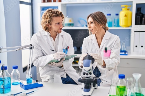 Man and woman wearing scientist uniform writing on notebook using microscope at laboratory