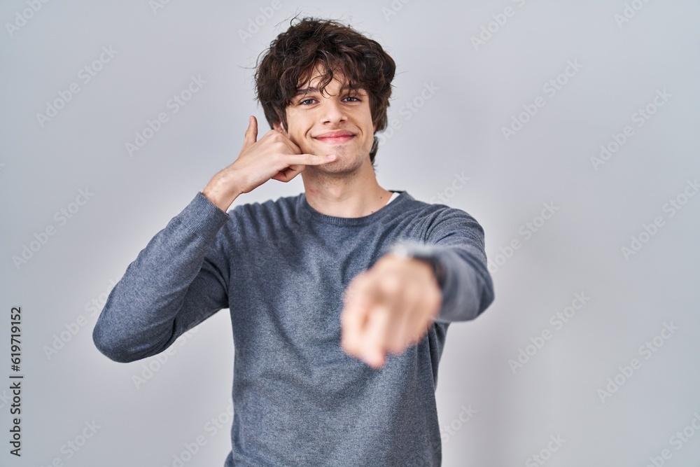 Young man standing over isolated background smiling doing talking on the telephone gesture and pointing to you. call me. Young man standing over isolated background smiling doing talking on the telephone gesture and pointing to you. call me.