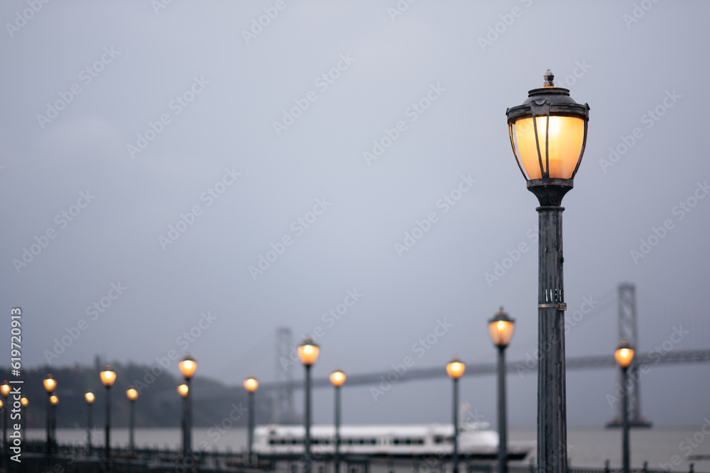 On a foggy day in San Francisco, a pier extends off into the distance, lined by antique lampposts with warm bulbs.