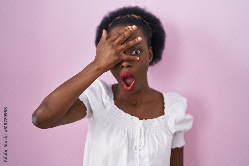 African woman with curly hair standing over pink background peeking in shock covering face and eyes with hand, looking through fingers with embarrassed expression. African woman with curly hair standing over pink background peeking in shock covering face and eyes with hand, looking through fingers with embarrassed expression.