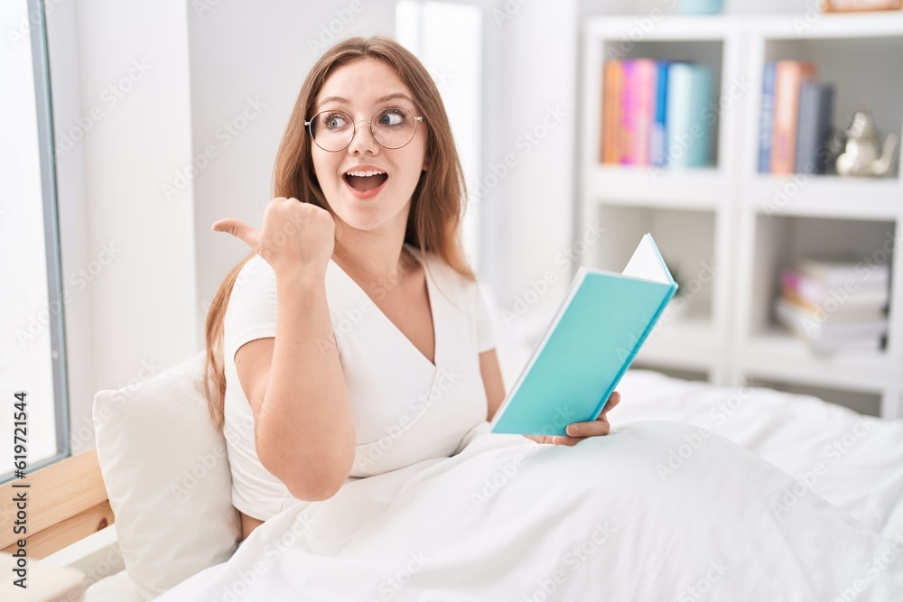 Young caucasian woman sitting on the bed at home reading a book pointing thumb up to the side smiling happy with open mouth Young caucasian woman sitting on the bed at home reading a book pointing thumb up to the side smiling happy with open mouth