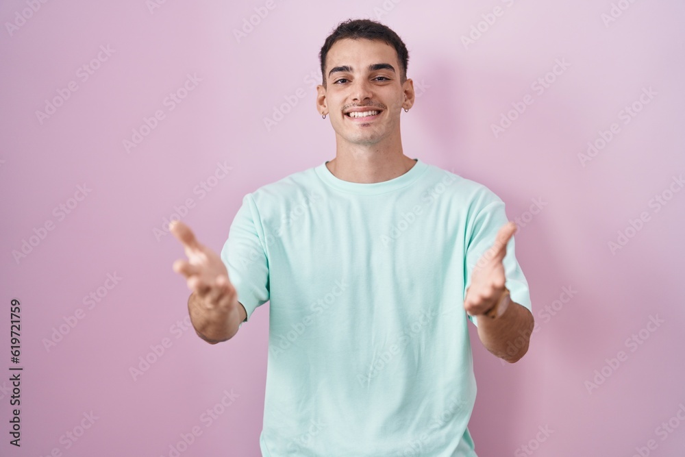 Handsome hispanic man standing over pink background smiling cheerful offering hands giving assistance and acceptance. Handsome hispanic man standing over pink background smiling cheerful offering hands giving assistance and acceptance.