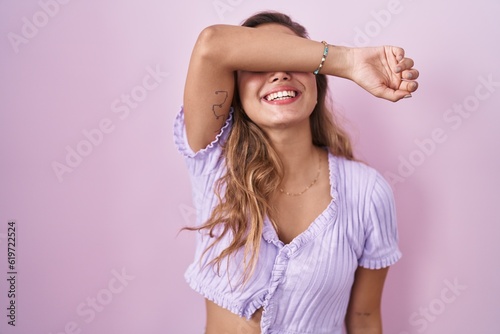 Young hispanic woman standing over pink background covering eyes with arm smiling cheerful and funny. blind concept.