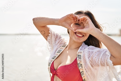 Young beautiful hispanic woman tourist wearing bikini doing heart gesture with hands at beach