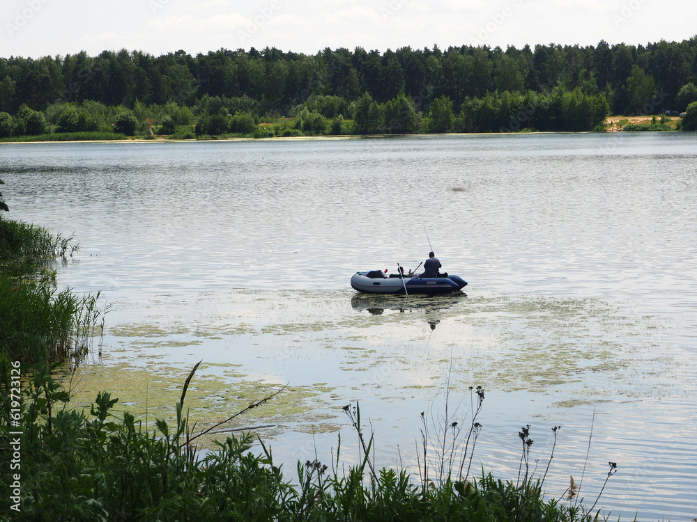A fisherman sitting in a rubber boat, the landscape of the lake on a summer day. Active sports and recreation on the pond.