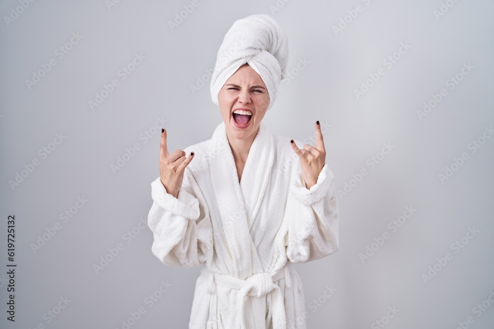 Blonde caucasian woman wearing bathrobe shouting with crazy expression doing rock symbol with hands up. music star. heavy music concept. Blonde caucasian woman wearing bathrobe shouting with crazy expression doing rock symbol with hands up. music star. heavy music concept.