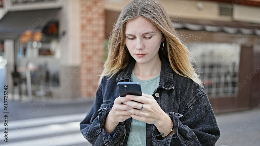 Young blonde woman using smartphone with serious face at coffee shop terrace