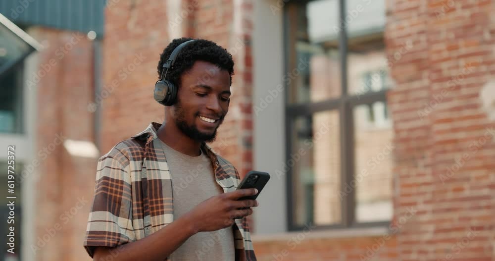Joyful Black millennial guy listening to music in headphones while using phone. 