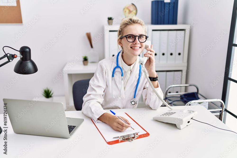 Young blonde woman doctor talking on telephone writing on document at clinic