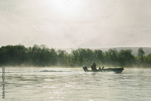 A fisherman with two dogs in a boat sails down the river early in the morning. Morning fog, sun in haze. High quality photo