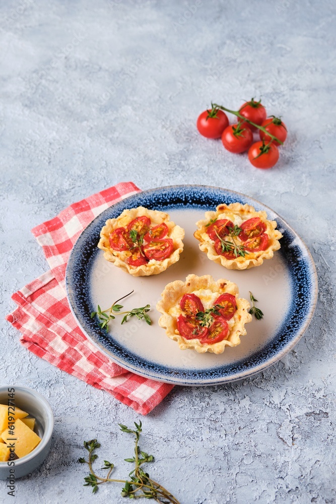 Unsweetened shortbread tartlets with feta cheese, cherry tomatoes and herbs on a blue plate on a light concrete background.