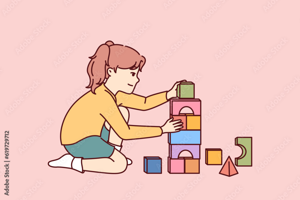 Little girl plays sitting on floor and builds toy bricks tower for concept of educational games for children. Child uses plastic or wooden toy bricks enjoying creation of pyramids.