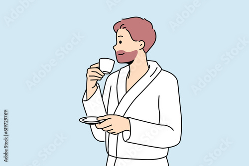 Man in bathrobe drinks coffee enjoying hot tart drink after waking up in morning. Successful young man with tea having rest in hotel or using services of spa center during vacation or weekend