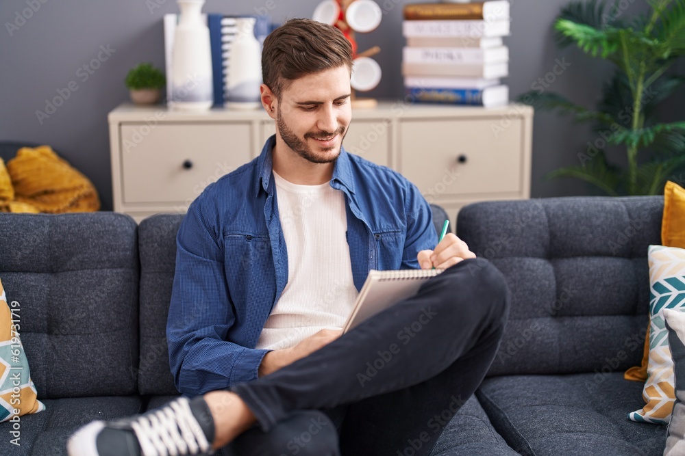 Young caucasian man writing on notebook sitting on sofa at home Young caucasian man writing on notebook sitting on sofa at home