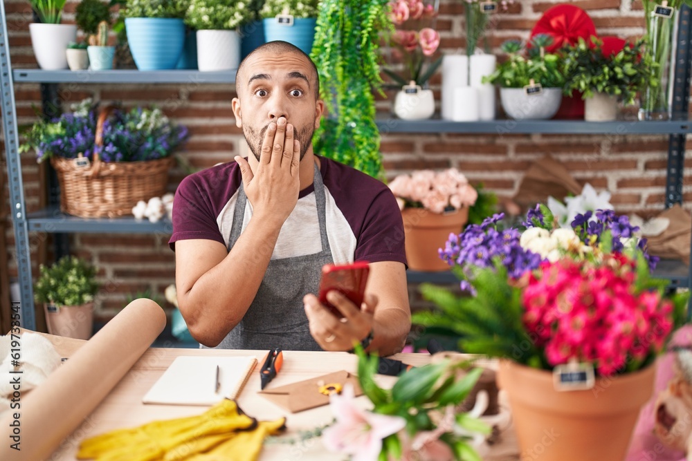 Hispanic man with beard working at florist shop with smartphone covering mouth with hand, shocked and afraid for mistake. surprised expression Hispanic man with beard working at florist shop with smartphone covering mouth with hand, shocked and afraid for mistake. surprised expression