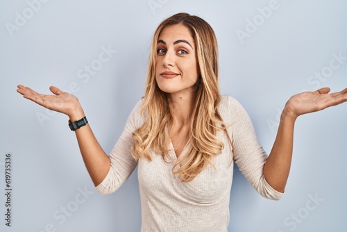Young blonde woman standing over isolated background clueless and confused expression with arms and hands raised. doubt concept.