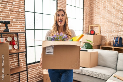 Young blonde woman moving to a new home holding cardboard box angry and mad screaming frustrated and furious, shouting with anger looking up. © Krakenimages.com