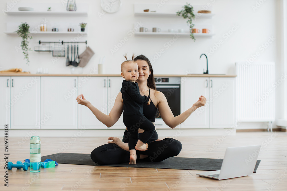 Energetic infant girl joining lady during her achieving cross-legged position due to online yoga class on device. Happy mother promoting daughter's sense of calmness by meditating together at home. Energetic infant girl joining lady during her achieving cross-legged position due to online yoga class on device. Happy mother promoting daughter's sense of calmness by meditating together at home.