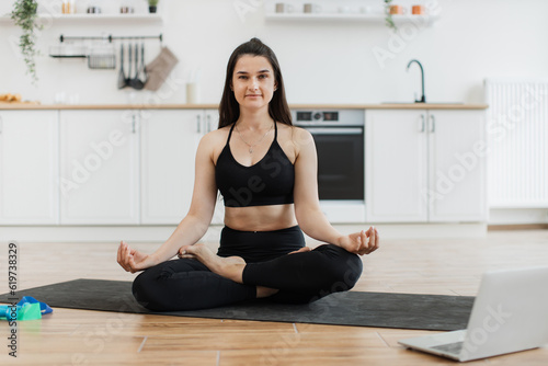 Portrait of young healthy female in yoga clothing breathing in lotus position with gyan mudra on rubber mat at home. Peaceful caucasian person encouraging active lifestyle with online classes.