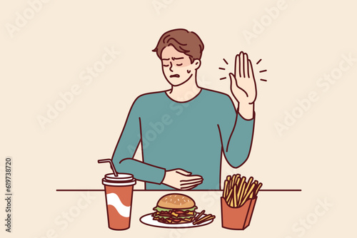 Man suffering from gastritis refuses fast food that causes pain in abdomen on advice of nutritionist. Guy near table with junk lunch shows stop gesture due to gastritis caused by eating hamburgers