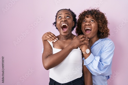 Two african women standing over pink background angry and mad screaming frustrated and furious, shouting with anger looking up.