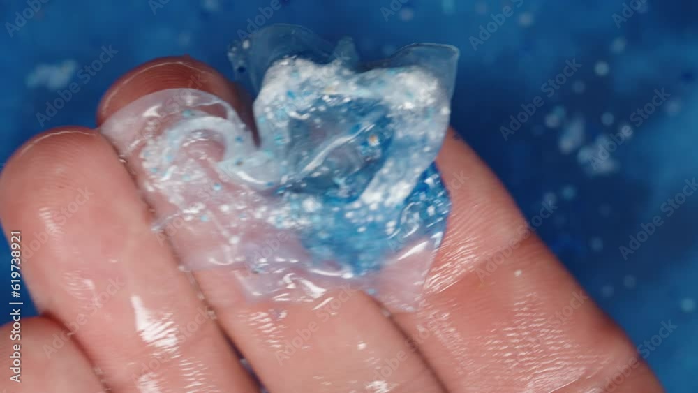 A soluble packet with blue and white powder, for a dishwasher, being held in my hand against a background of blue water where one has already dissolved. Close-up shot. A soluble packet with blue and white powder, for a dishwasher, being held in my hand against a background of blue water where one has already dissolved. Close-up shot.