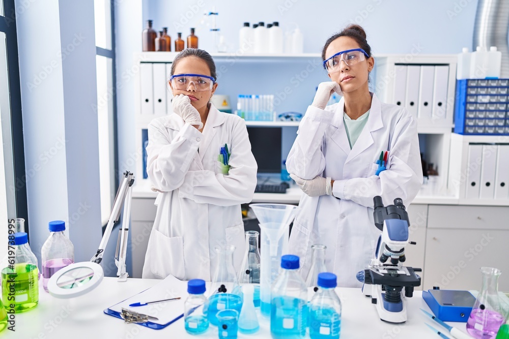 Young mother and daughter at scientist laboratory serious face thinking about question with hand on chin, thoughtful about confusing idea Young mother and daughter at scientist laboratory serious face thinking about question with hand on chin, thoughtful about confusing idea