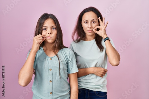 Young mother and daughter standing over pink background mouth and lips shut as zip with fingers. secret and silent, taboo talking