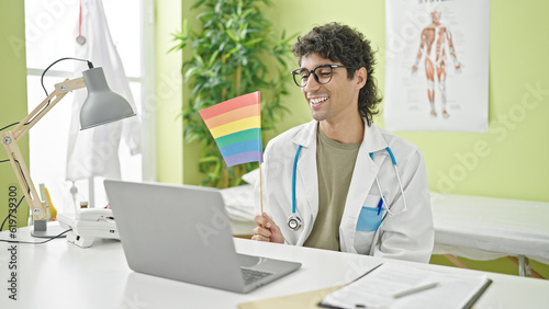 Young hispanic man doctor having video call holding rainbow flag at clinic