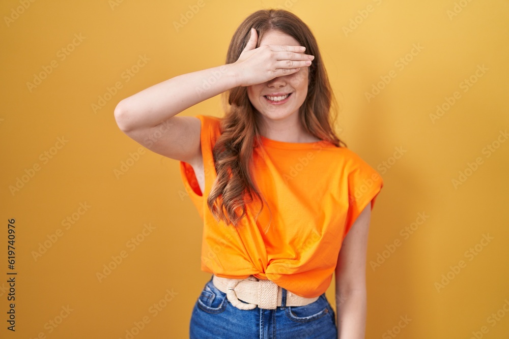 Caucasian woman standing over yellow background smiling and laughing with hand on face covering eyes for surprise. blind concept. Caucasian woman standing over yellow background smiling and laughing with hand on face covering eyes for surprise. blind concept.