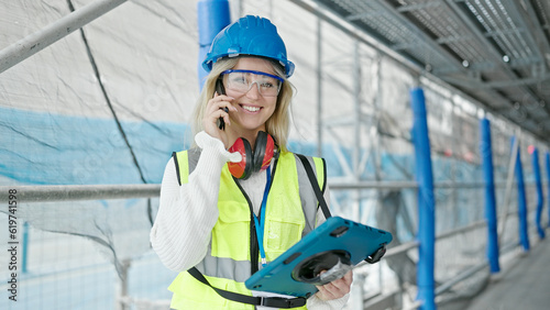 Young blonde woman architect talking on smartphone using touchpad at street