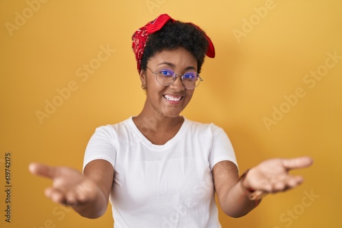 Young african american woman standing over yellow background smiling cheerful offering hands giving assistance and acceptance.