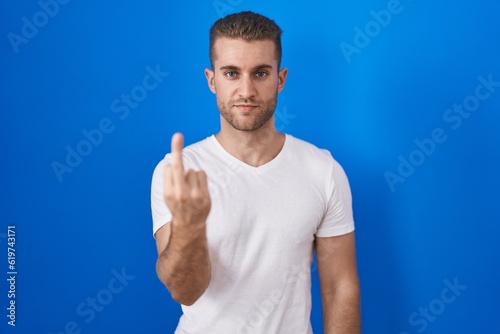 Young caucasian man standing over blue background showing middle finger, impolite and rude fuck off expression