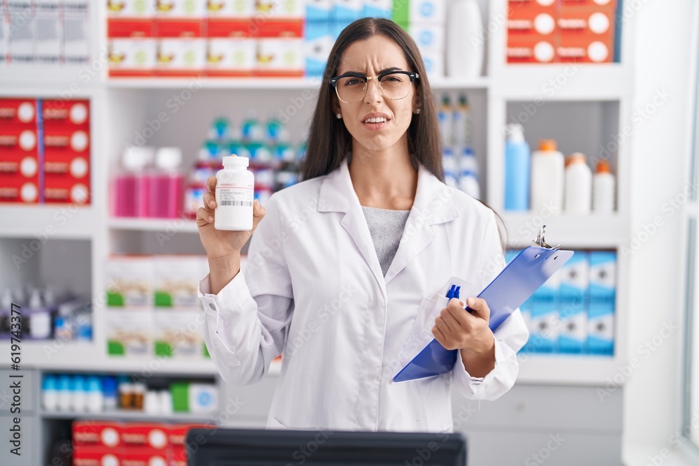 Young brunette woman working at pharmacy drugstore holding pills clueless and confused expression. doubt concept.