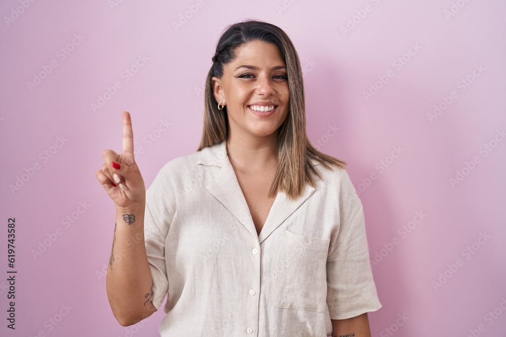 Blonde woman standing over pink background showing and pointing up with finger number one while smiling confident and happy. Blonde woman standing over pink background showing and pointing up with finger number one while smiling confident and happy.