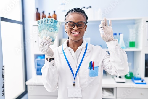 African woman with braids working at scientist laboratory holding money winking looking at the camera with sexy expression, cheerful and happy face.