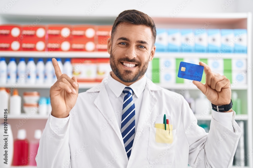 Handsome hispanic man working at pharmacy drugstore holding credit card smiling happy pointing with hand and finger to the side Handsome hispanic man working at pharmacy drugstore holding credit card smiling happy pointing with hand and finger to the side