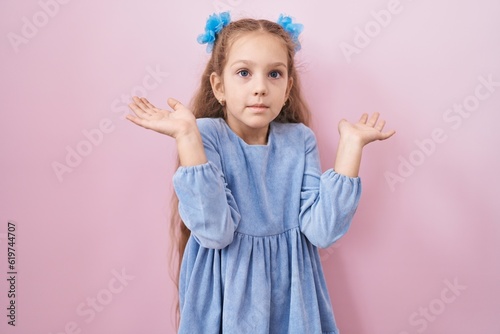 Young little girl standing over pink background clueless and confused expression with arms and hands raised. doubt concept.