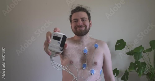 Young man with EKG heart monitor smiles and holds device towards camera 