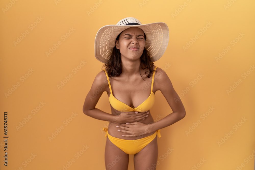 Young hispanic woman wearing bikini and summer hat with hand on stomach because indigestion, painful illness feeling unwell. ache concept. Young hispanic woman wearing bikini and summer hat with hand on stomach because indigestion, painful illness feeling unwell. ache concept.