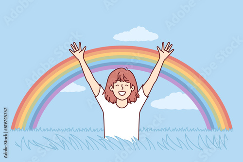 Happy girl rejoices at rainbow appeared in blue sky after rain, and runs across field with tall grass. Child teenager with smile raises hands enjoying spring weather and beautiful rainbow.