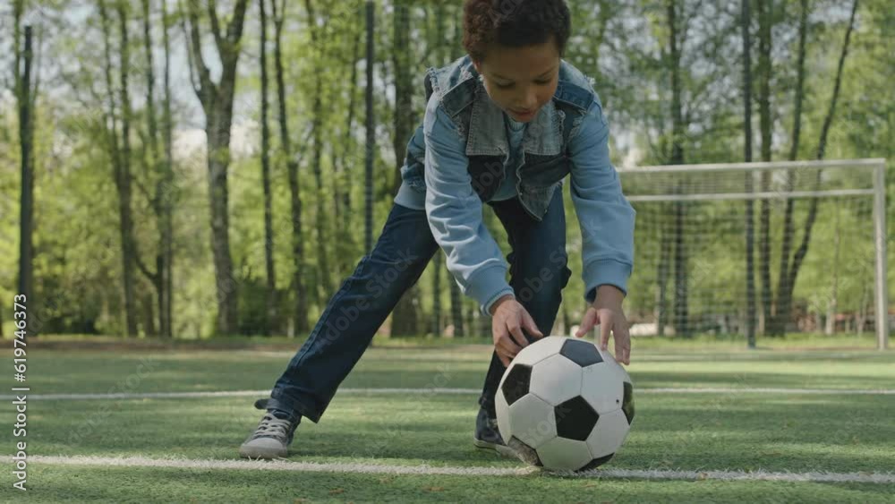 Cheerful African American boy walking on soccer field in park, putting ball on grass and preparing to kick it Cheerful African American boy walking on soccer field in park, putting ball on grass and preparing to kick it