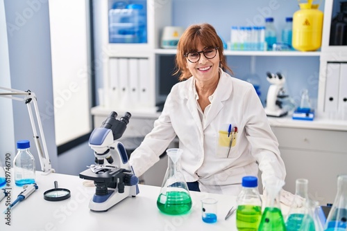 Middle age woman scientist smiling confident sitting on table at laboratory